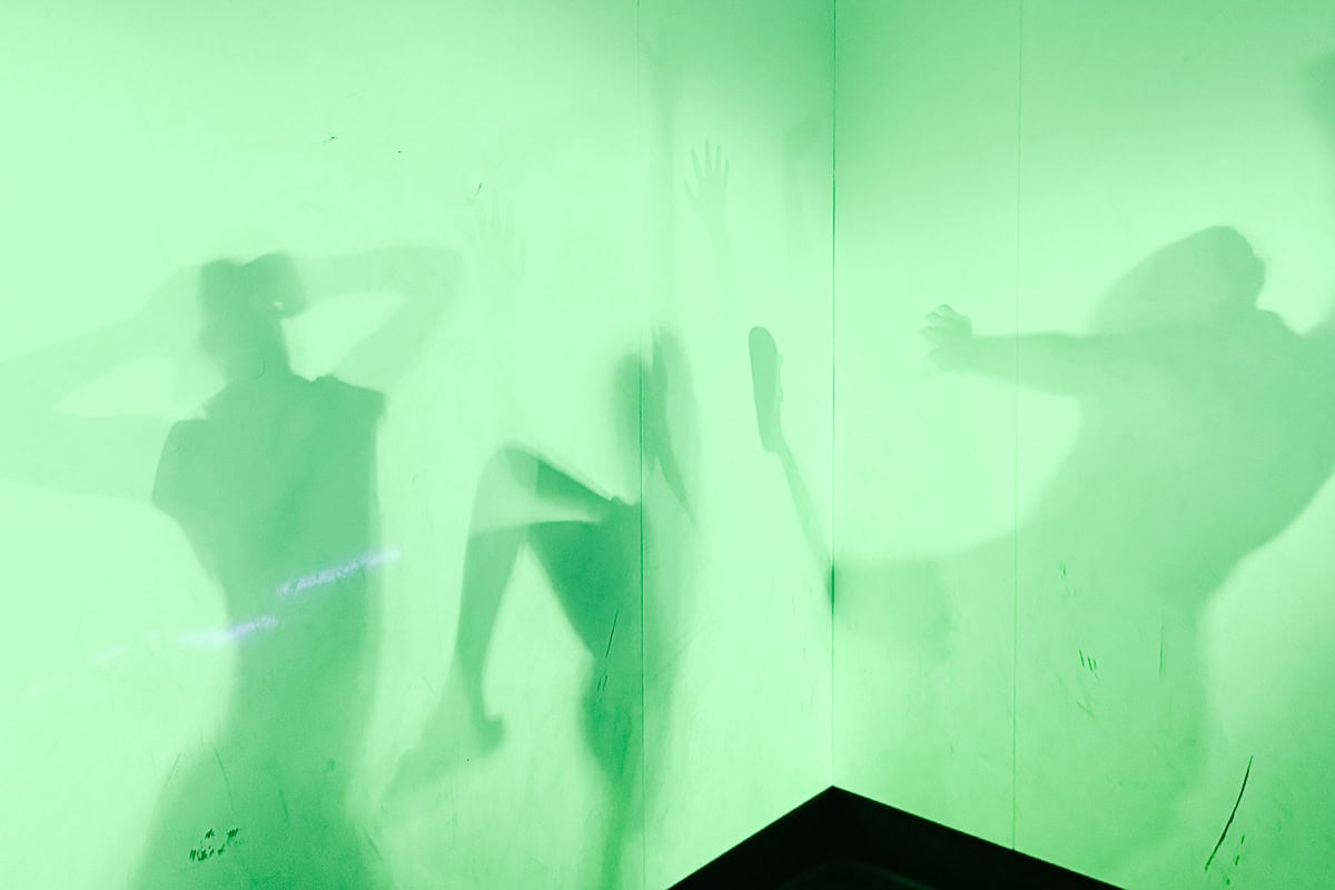 Green-lit room with three human shadows on the walls, each in a different dynamic pose—an immersive scene reminiscent of the creative attractions found among the things to do in Branson.