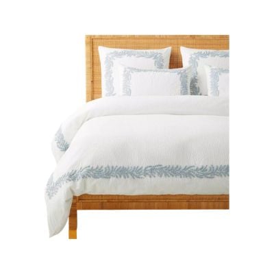 A neatly made bed with a light wood headboard, white bedding, and matching pillows featuring light blue embroidered borders and leaf patterns offers a chic serena and lily look for less.