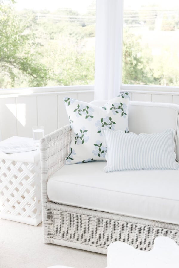White wicker sofa with two decorative pillows, one with a leaf pattern and one striped, beside a white side table and a glass of water in a bright, airy room—perfect for relaxing or upgrading your space this Prime Day.