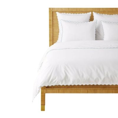 A wooden bed frame with a cane headboard is shown, topped with white bedding and pillows featuring a subtle blue trim, offering a beautiful Serena and Lily look for less.