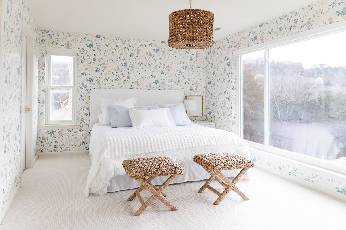 Bright bedroom with floral wallpaper, a white bed with light blue and white pillows, two woven stools at the foot for a Serena and Lily look for less, large window, and a woven pendant light fixture.
