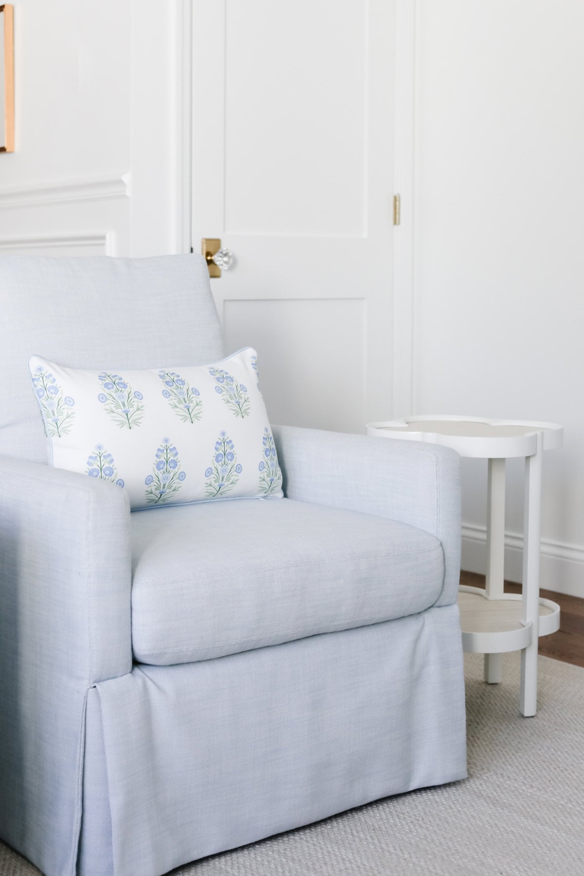 Light blue armchair with a floral pillow sits next to a small white side table in a bright, minimalist room with white walls and a closed door, creating a serena and lily look for less.