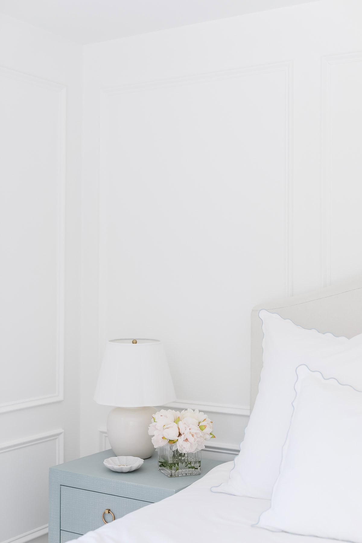 A light blue nightstand with a white lamp, a small bowl, and a vase of pale flowers is beside a bed with white scalloped-edge bedding in a white room, capturing the Serena and Lily look for less.