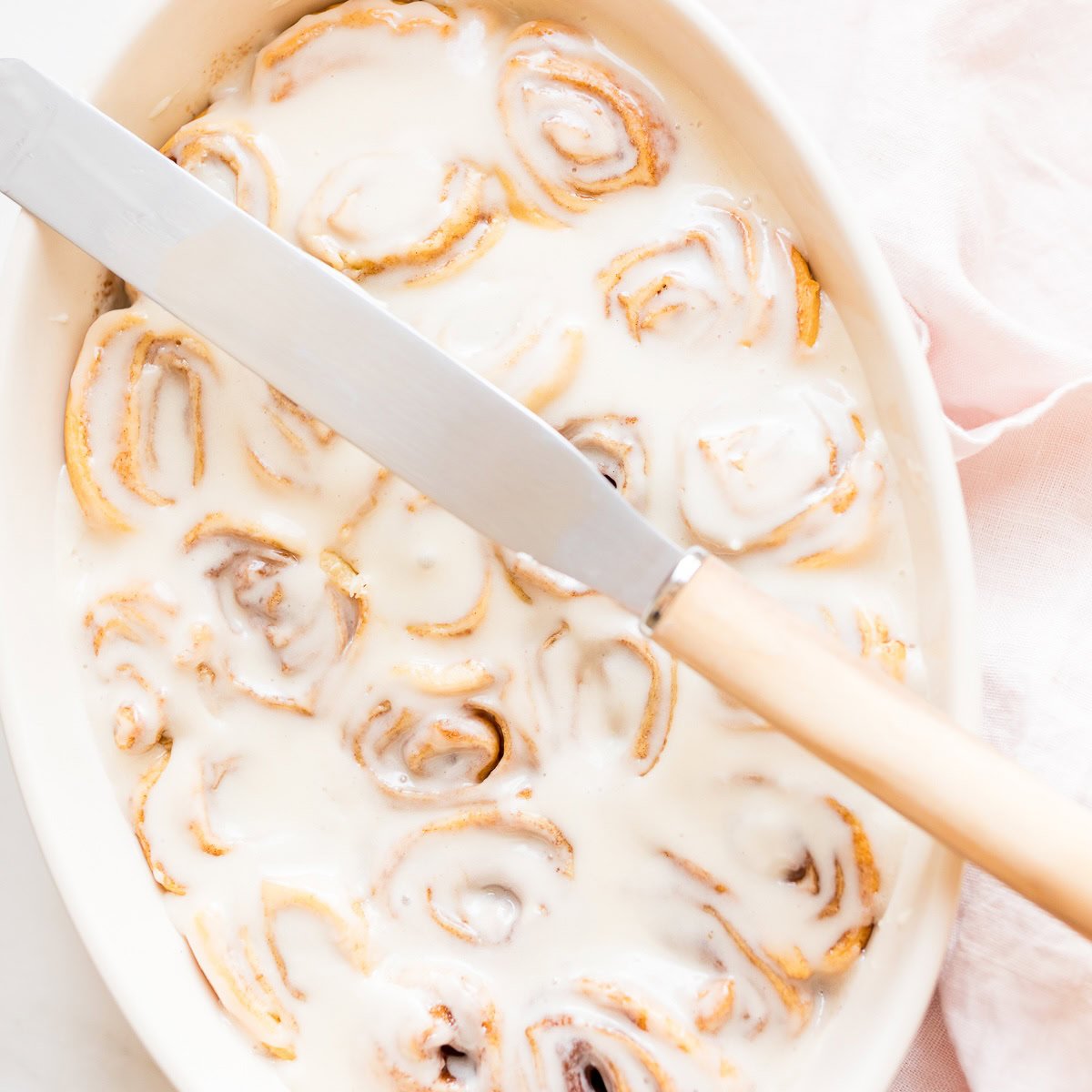 A dish of cinnamon rolls covered in white icing with a large knife resting on top, next to a light pink cloth.