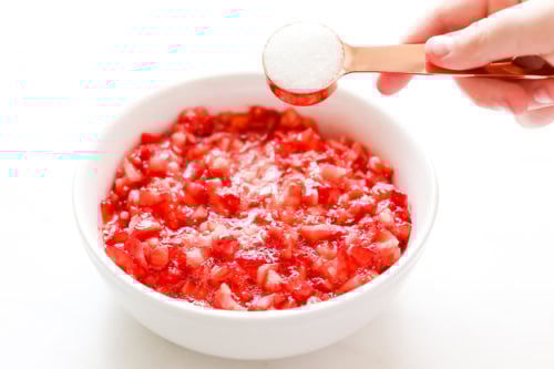 A hand holds a measuring spoon of sugar above a bowl of chopped strawberries.