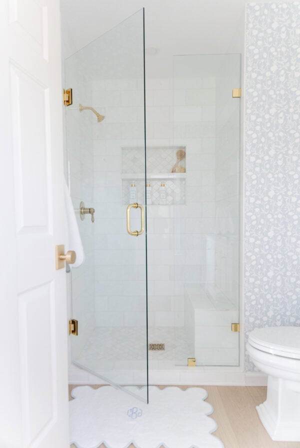 Bright bathroom with a glass shower enclosure, gold fixtures, white tile, a patterned wall, and a scalloped white bath mat on a light wood floor.