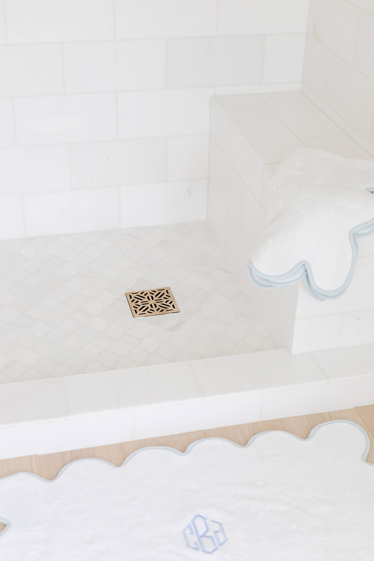 White tiled shower with a decorative gold drain cover, a built-in bench, and white towels with blue edging placed on the bench and floor.