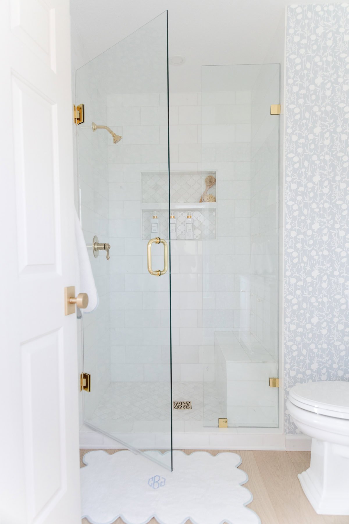 Bright bathroom with a glass shower enclosure, gold fixtures, white tile, a patterned wall, and a scalloped white bath mat on a light wood floor.
