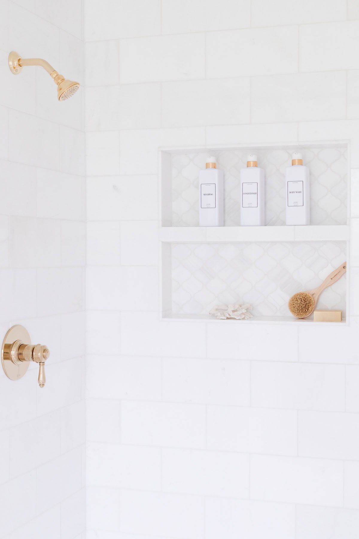 A minimalist white shower with gold fixtures, built-in shelves holding three labeled bottles, a brush, and natural sponges.