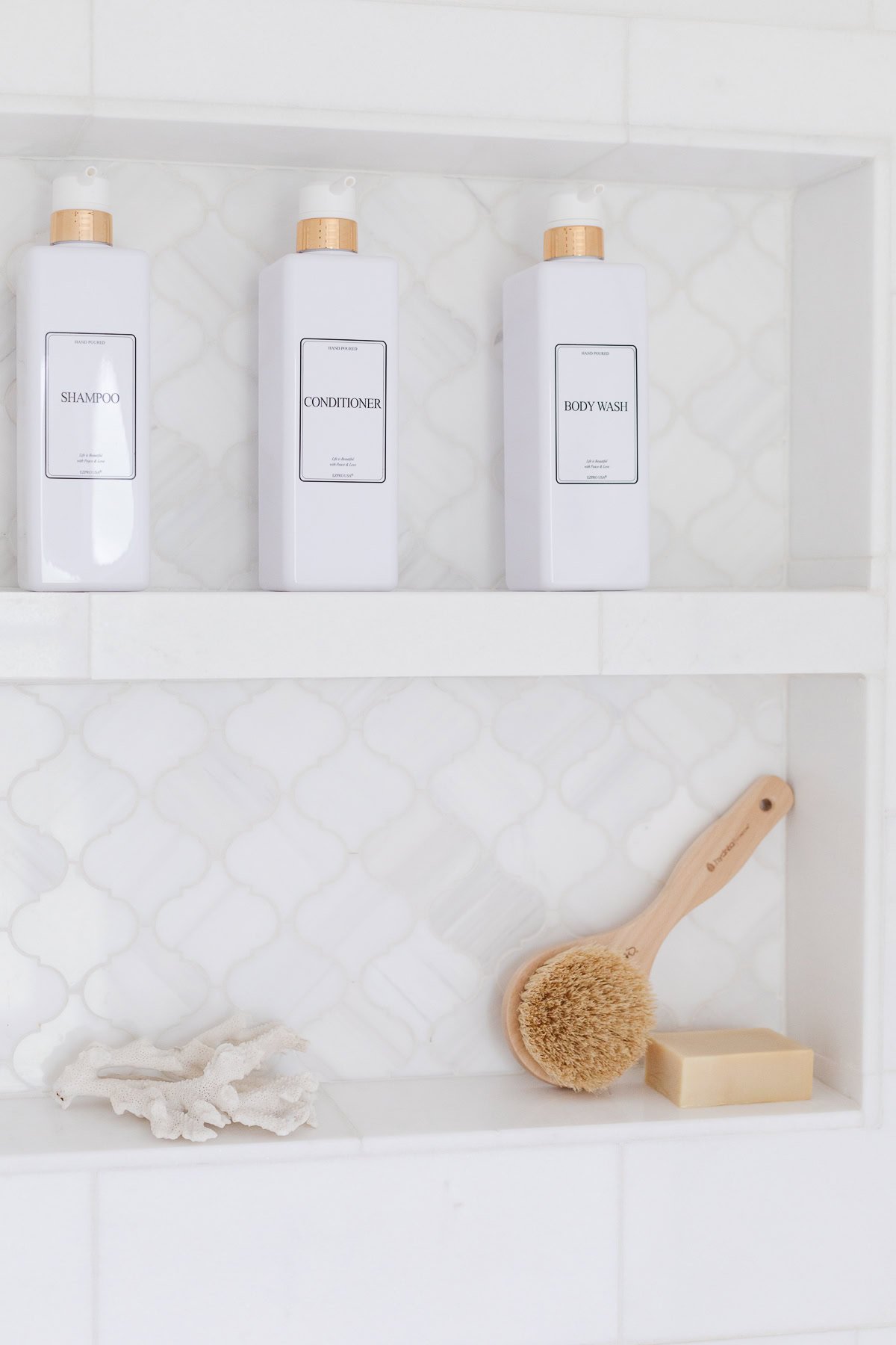 White bathroom shelves with bottles labeled shampoo, conditioner, and body wash on the top shelf, and a brush, bar of soap, and sponge on the lower shelf.