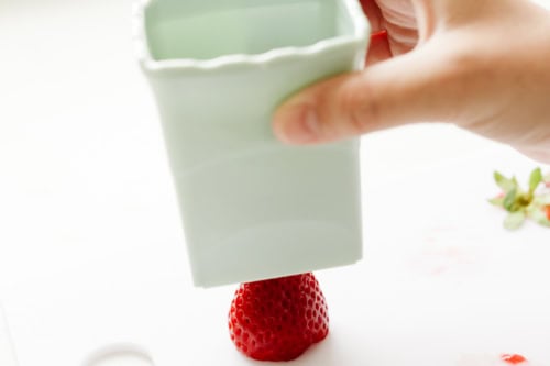 A hand uses a light green plastic container to hull a strawberry, with the strawberry positioned on a white surface.