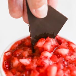 A hand dips a piece of dark chocolate into a bowl of diced strawberries in syrup.