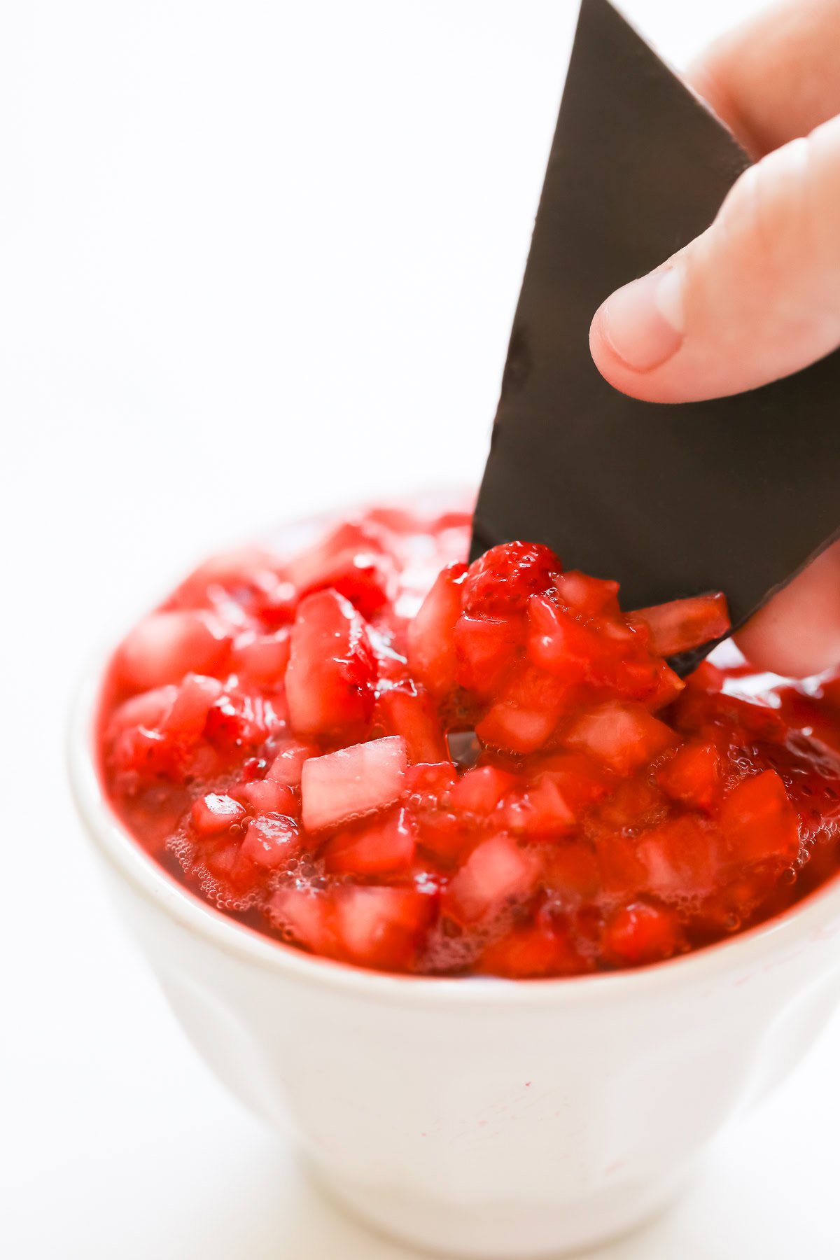 A hand dips a piece of dark chocolate into a white bowl filled with chopped strawberries and sauce.