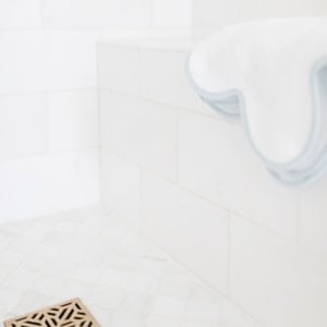 A white towel with blue trim is draped over the edge of a white tiled shower bench above a tiled floor with a metal drain.