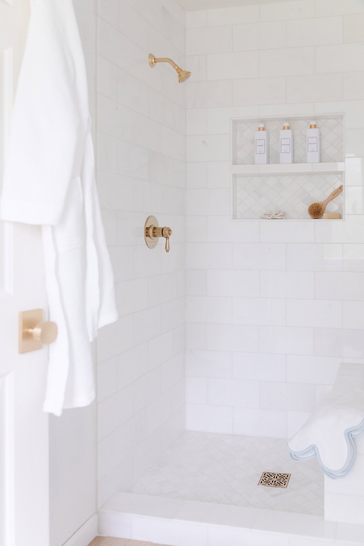 A white tiled shower with gold fixtures, built-in shelves holding bottles and a brush, and a white robe hanging on the door.