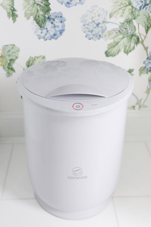 A white Conffier diaper pail with a closed lid stands on a white tile floor in front of floral wallpaper.