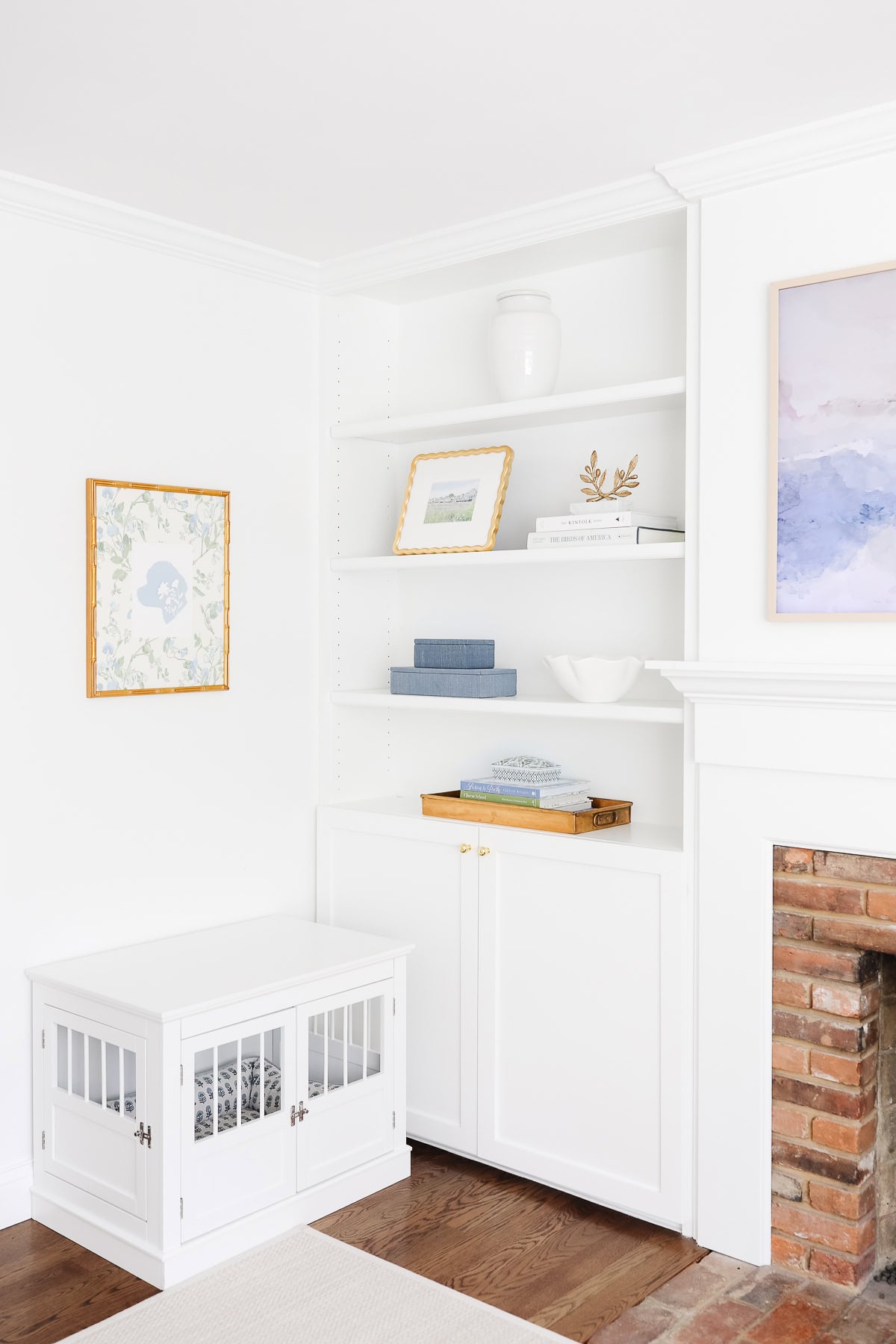 A white built-in bookcase with shelves holding books and decor stands next to a fireplace; a white dog crate is placed beside it.