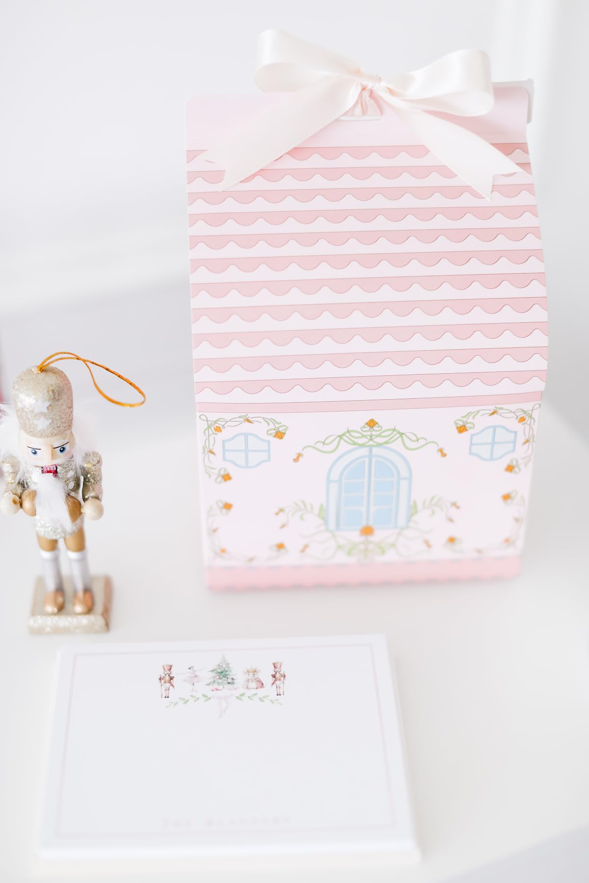 A decorative pink and white house-shaped box with a bow, a small nutcracker figurine, and a stack of notecards on a white surface—perfect for adding festive charm to your space this Prime Day.