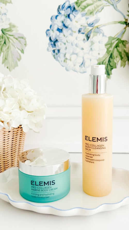 A jar of Elemis Pro-Collagen Marine Body Cream and a bottle of Elemis Pro-Collagen Bath & Shower Elixir sit on a tray beside white flowers and a floral wallpaper background.