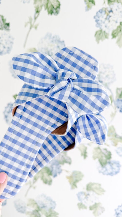 A pair of blue and white gingham sandals with large bows, held up against a floral patterned background.