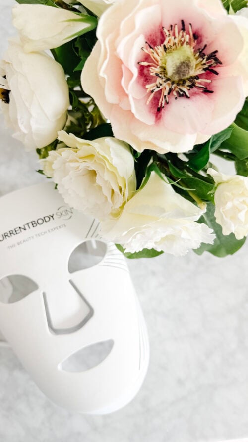 A white LED face mask and a bouquet of light pink and white flowers are arranged on a marble surface.