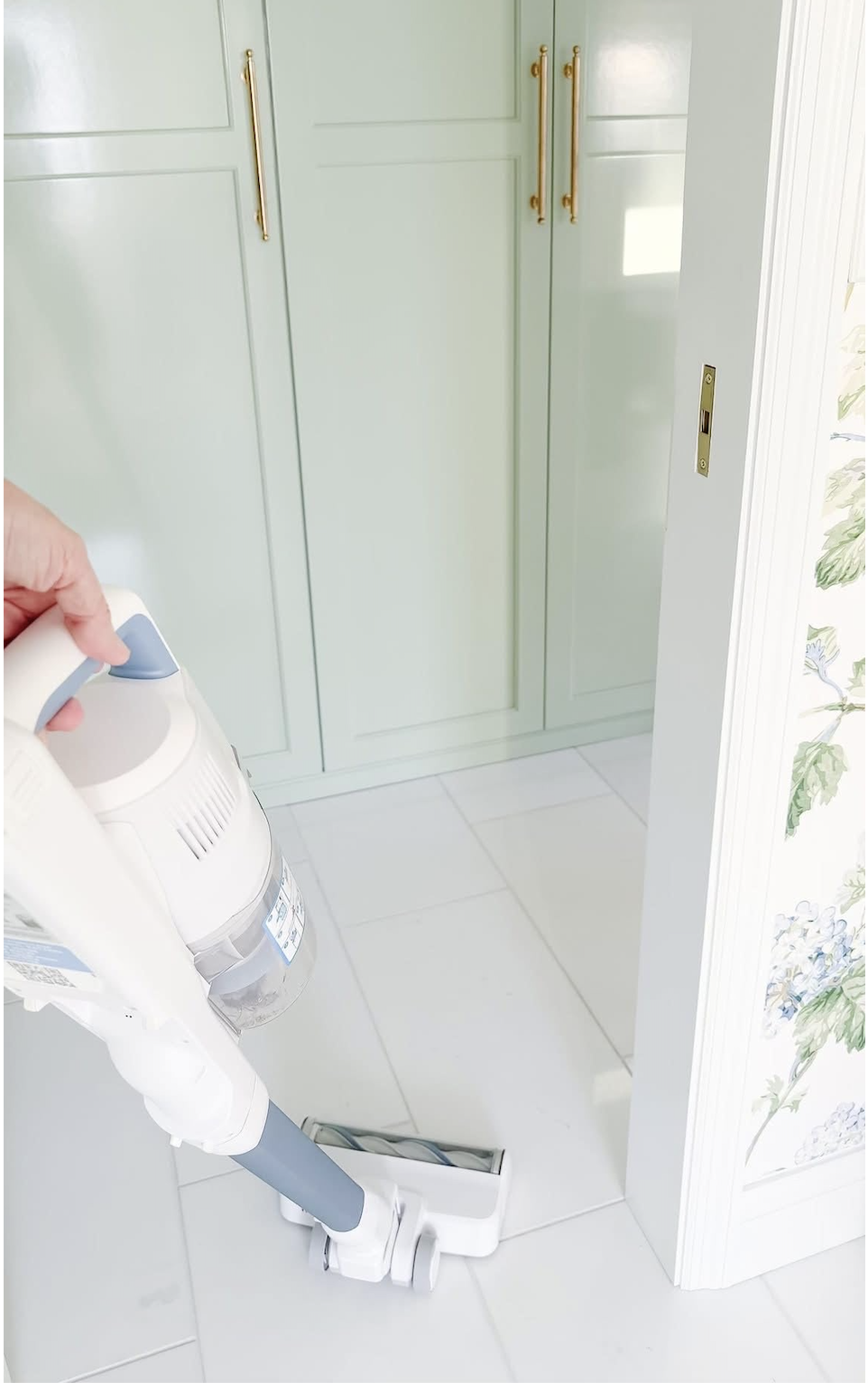 A person uses a white cordless vacuum cleaner on a white tile floor in front of light green cabinets with gold handles.