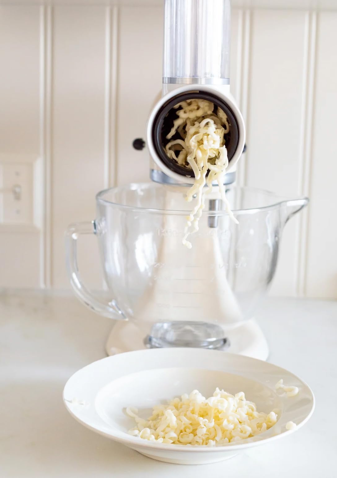 A stand mixer cheese grater attachment is shredding cheese into a glass bowl, with grated cheese falling into a white plate below.