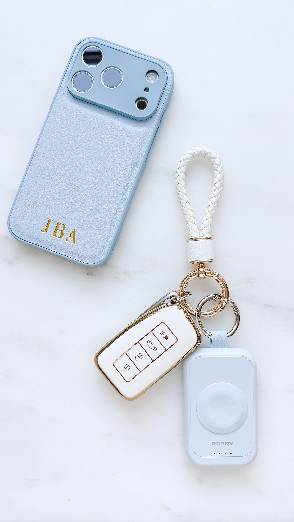 A light blue smartphone with "JBA" initials, a white and gold key fob, and a small light blue electronic device on a braided keychain, all on a white surface.