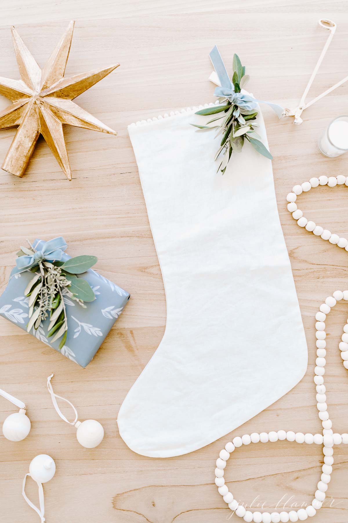 A white Christmas stocking with greenery sits on a light wooden surface, surrounded by a gold star, beads, white ornaments, a candle, and a wrapped gift—perfect for adding festive charm to your Prime Day holiday shopping haul.