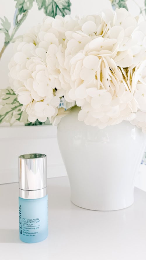 A blue bottle of Elemis Pro-Collagen moisturizer stands next to a white vase filled with white hydrangeas on a white surface.