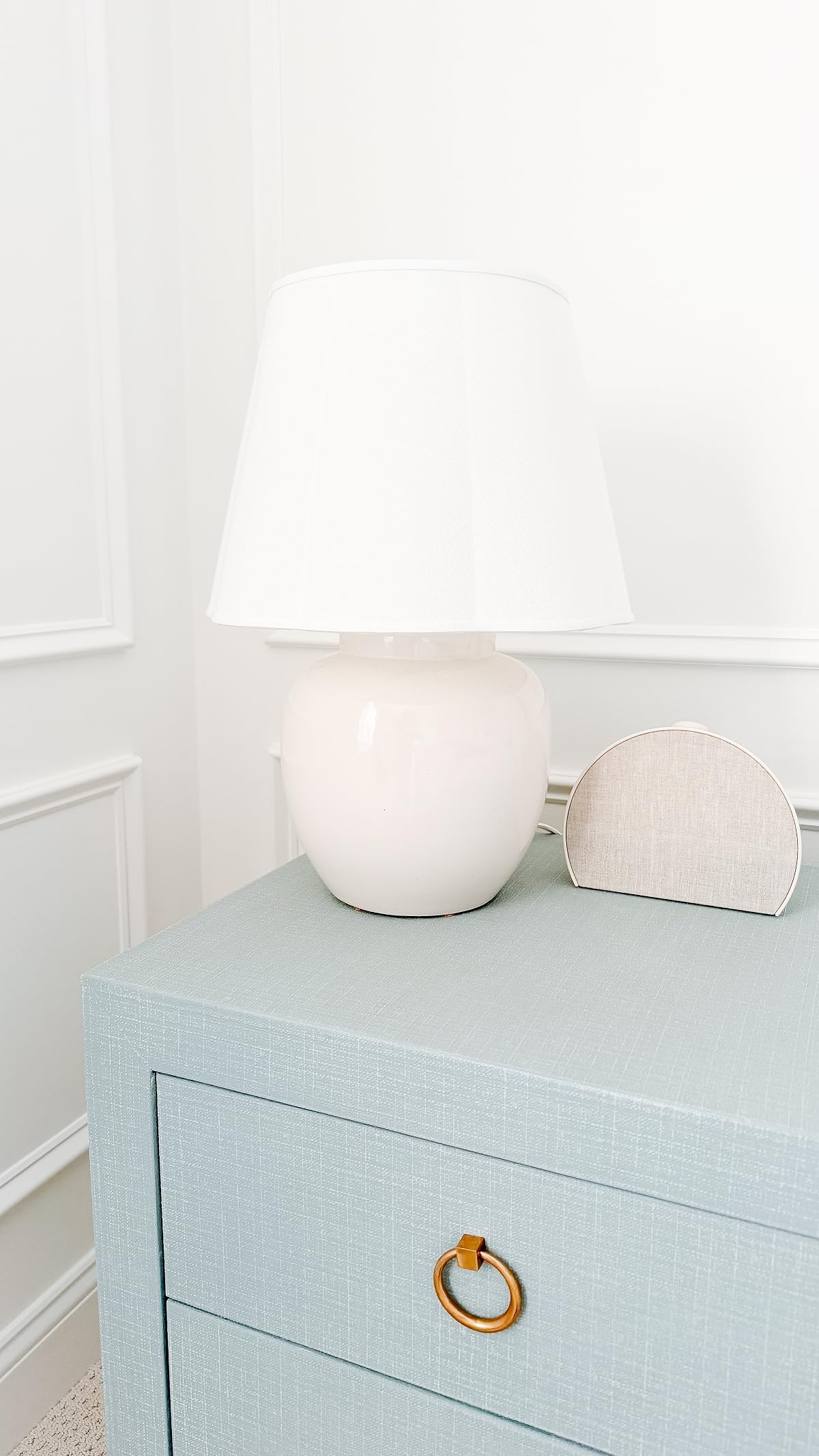 A white table lamp and a small beige speaker sit on top of a light blue dresser with a gold ring handle against a white wall.