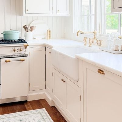 Bright kitchen with white cabinets, farmhouse sink, gold hardware, a stove with a pot, and a large window allowing natural light in.