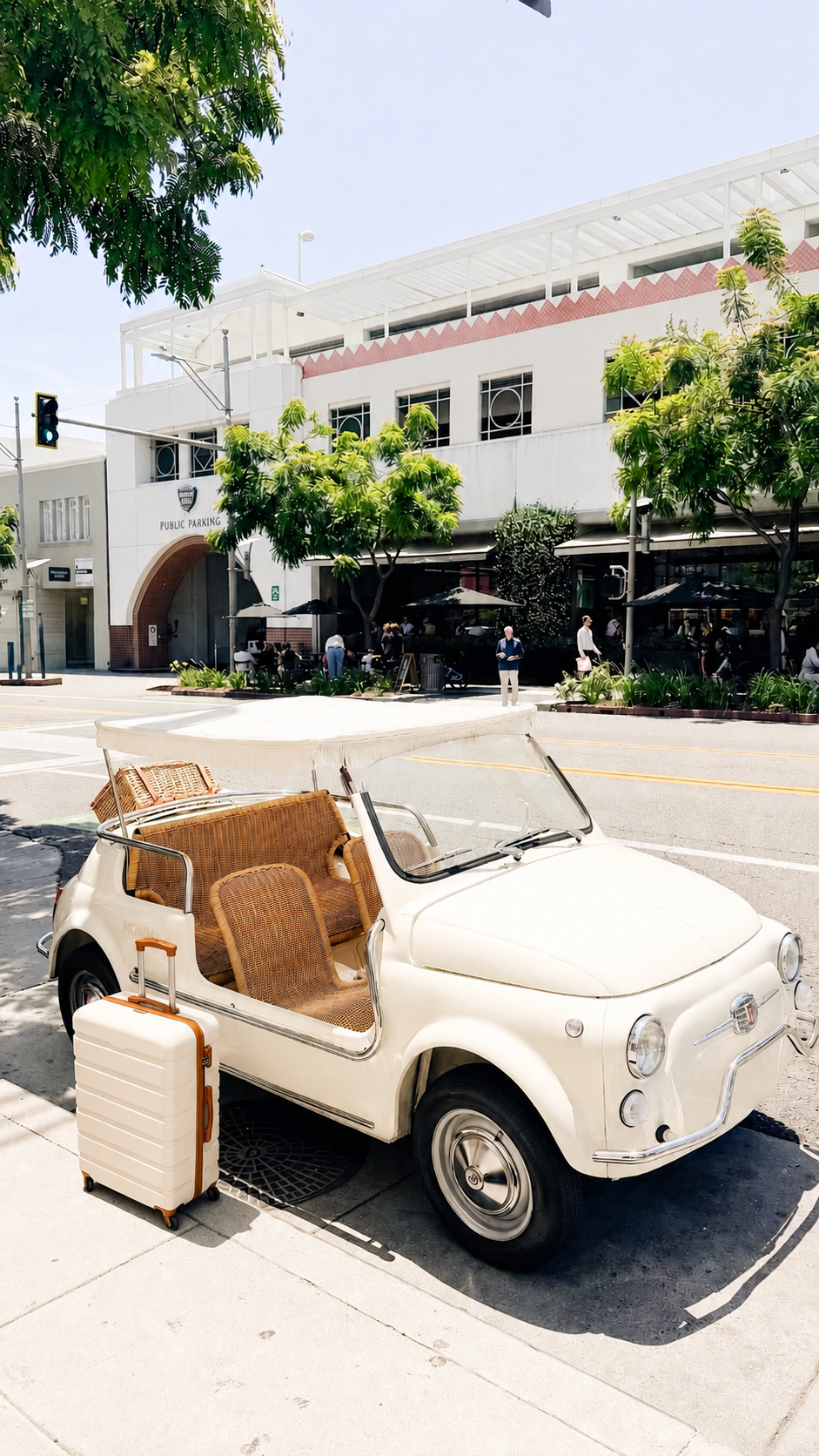 A small white vintage convertible with wicker seats is parked by the sidewalk, next to a beige suitcase, in front of a tree-lined building.