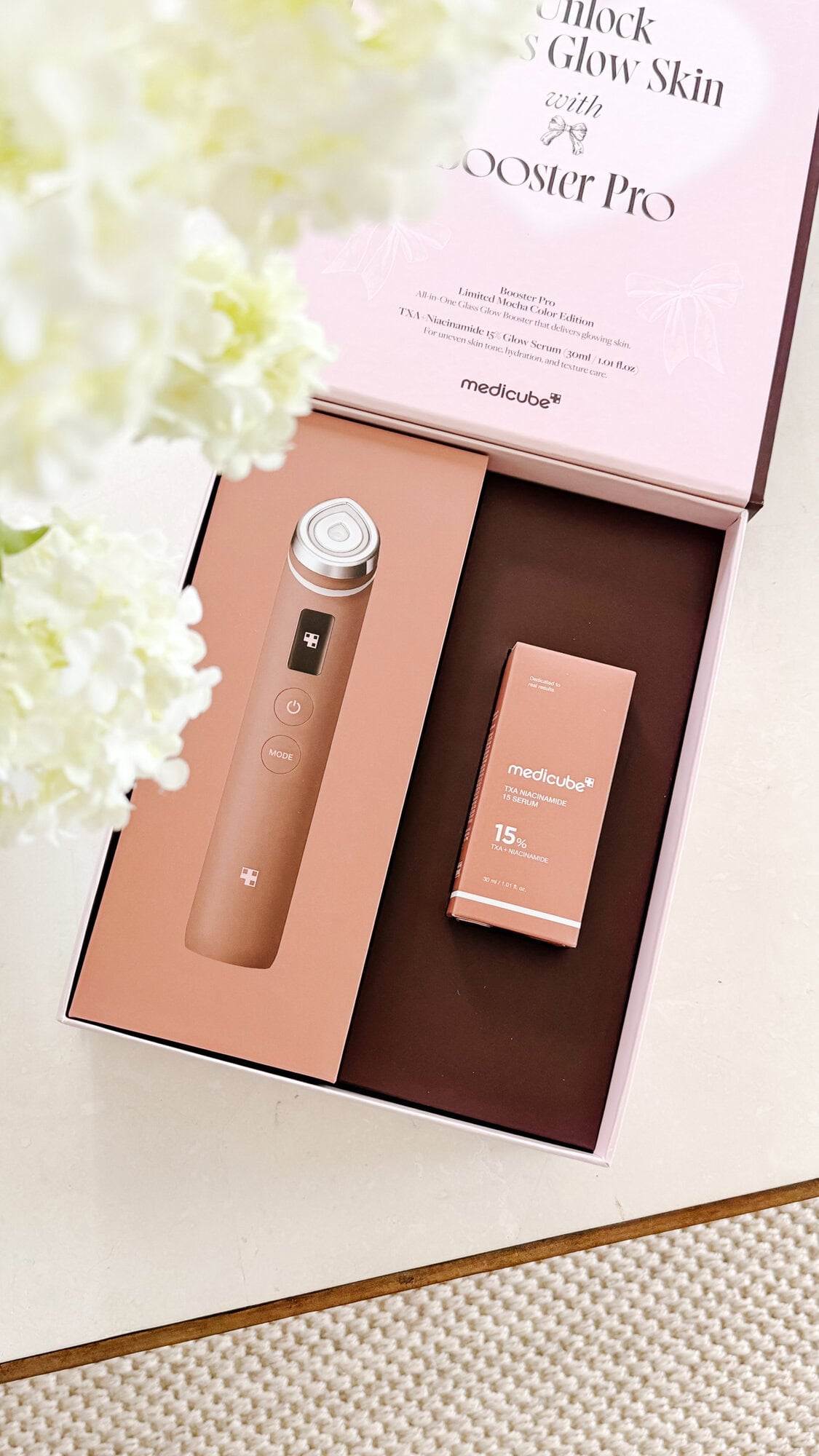 Open box with a Mediacube beauty device and a packaged skincare ampoule, placed on a light surface next to white flowers.
