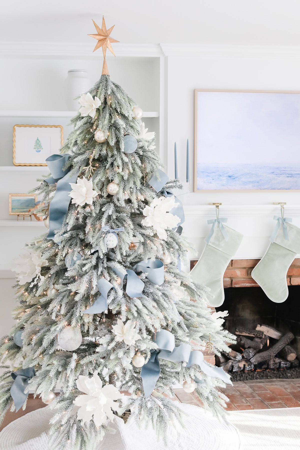 A flocked Christmas tree decorated with blue ribbons, white flowers, and ornaments, topped with a wooden star, standing beside a fireplace with two hanging stockings.