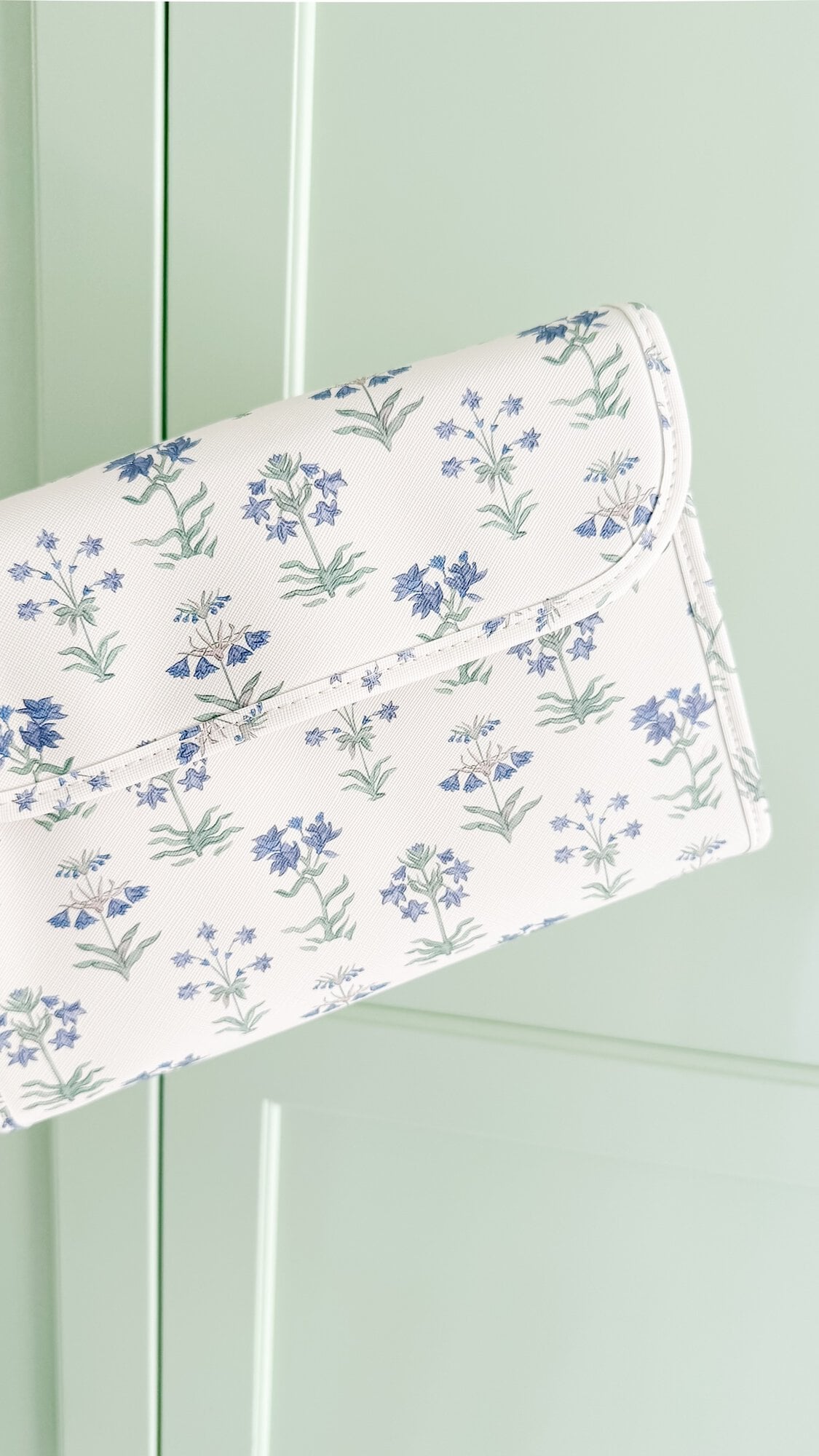 A white clutch purse with a blue and green floral pattern is displayed against a light green background.