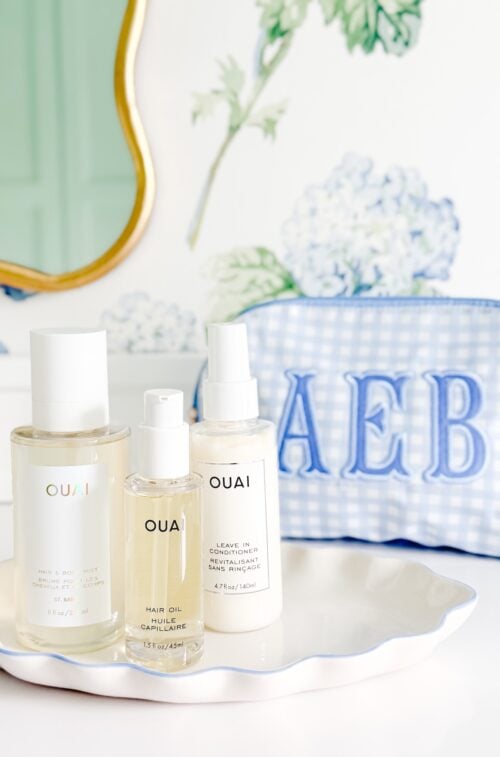 Three OUAI hair products—a hair and body mist, hair oil, and leave-in conditioner—on a scalloped white tray next to a blue gingham travel bag monogrammed with “AEB.”.