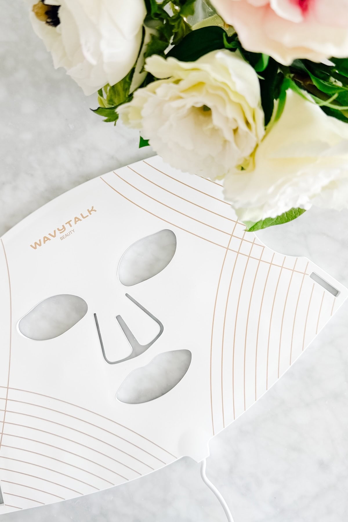 A white WavyTalk Beauty LED face mask next to a bouquet of white flowers on a marble surface.