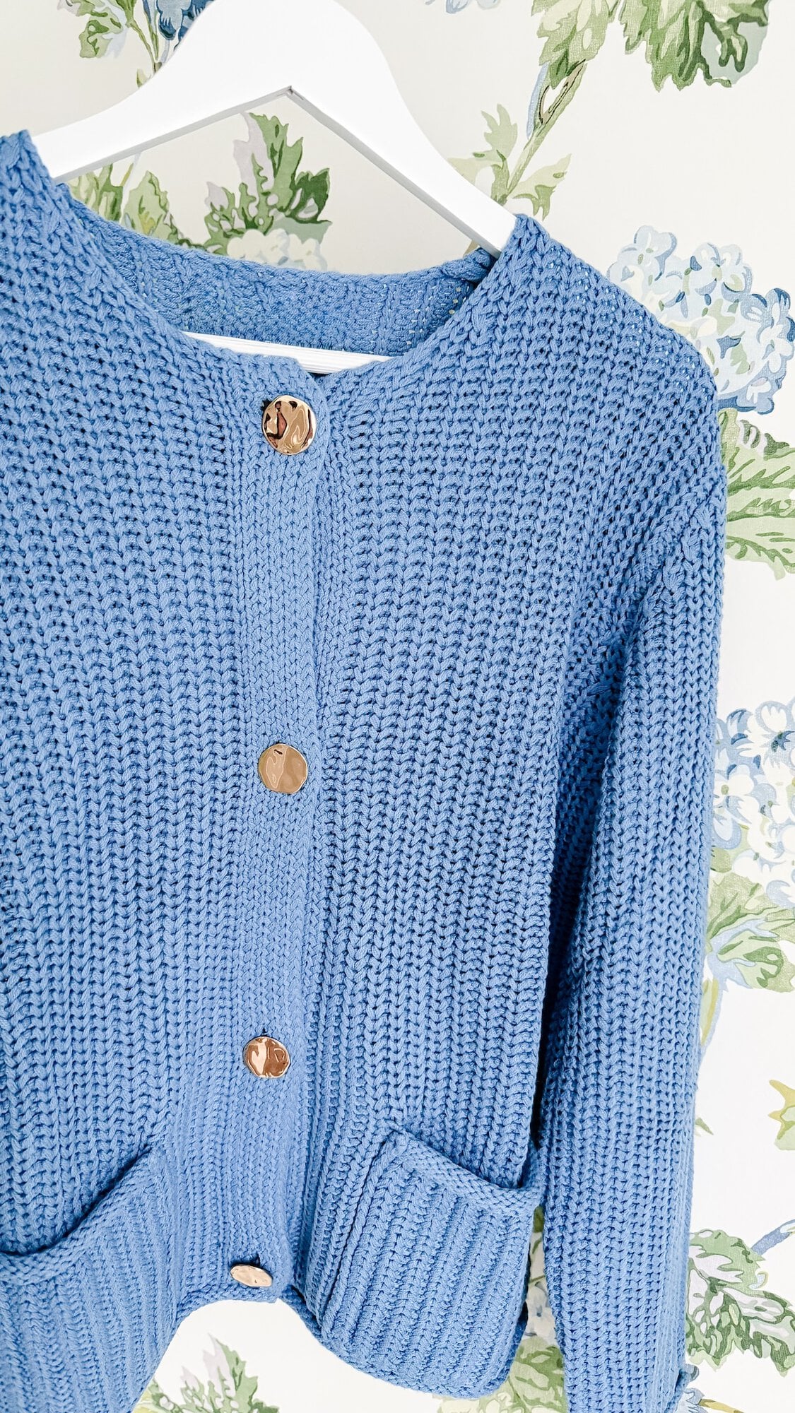 Blue knit cardigan with large gold buttons hanging on a white hanger.
