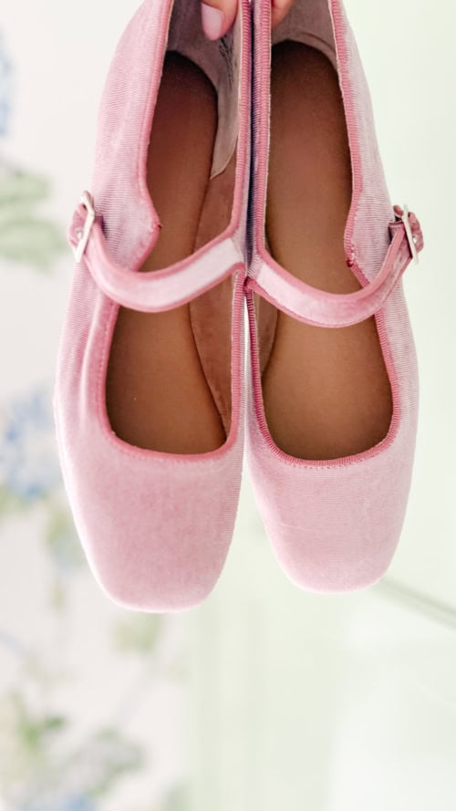 A pair of pink velvet Mary Jane shoes with a single strap, viewed from above against a soft, blurred background.