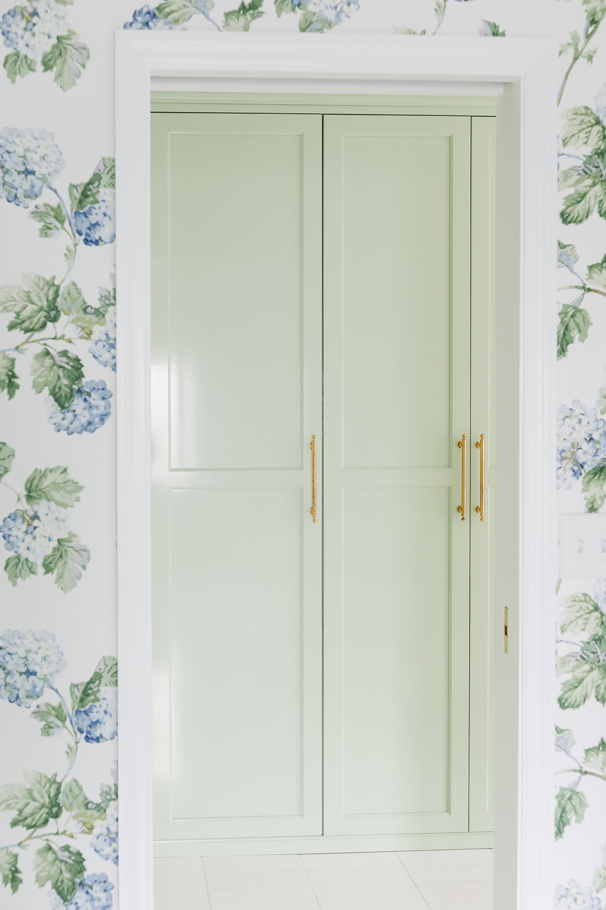 Light green double doors with gold handles are seen through a doorway, framed by floral wallpaper patterned with blue hydrangeas and green leaves against a Sherwin Williams Extra White background.