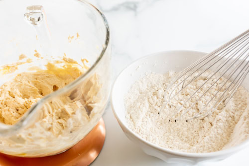A glass mixing bowl with creamed dough next to a white bowl containing flour and a metal whisk on a white countertop.