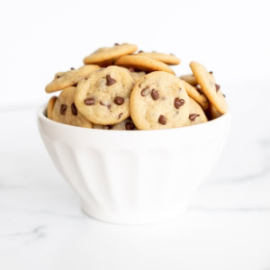 A white bowl filled with mini chocolate chip cookies sits on a white surface with a plain background.