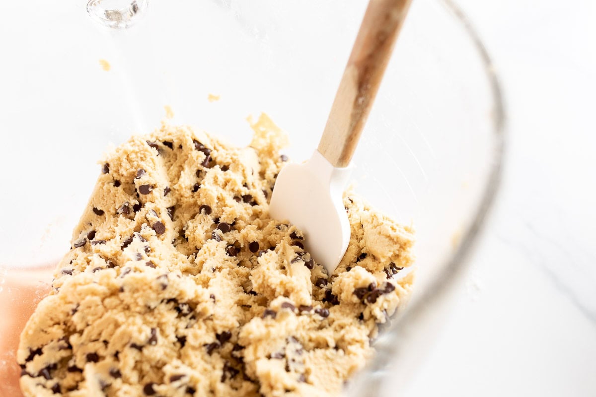 A glass mixing bowl containing chocolate chip cookie dough with a white spatula partially embedded in the mixture.