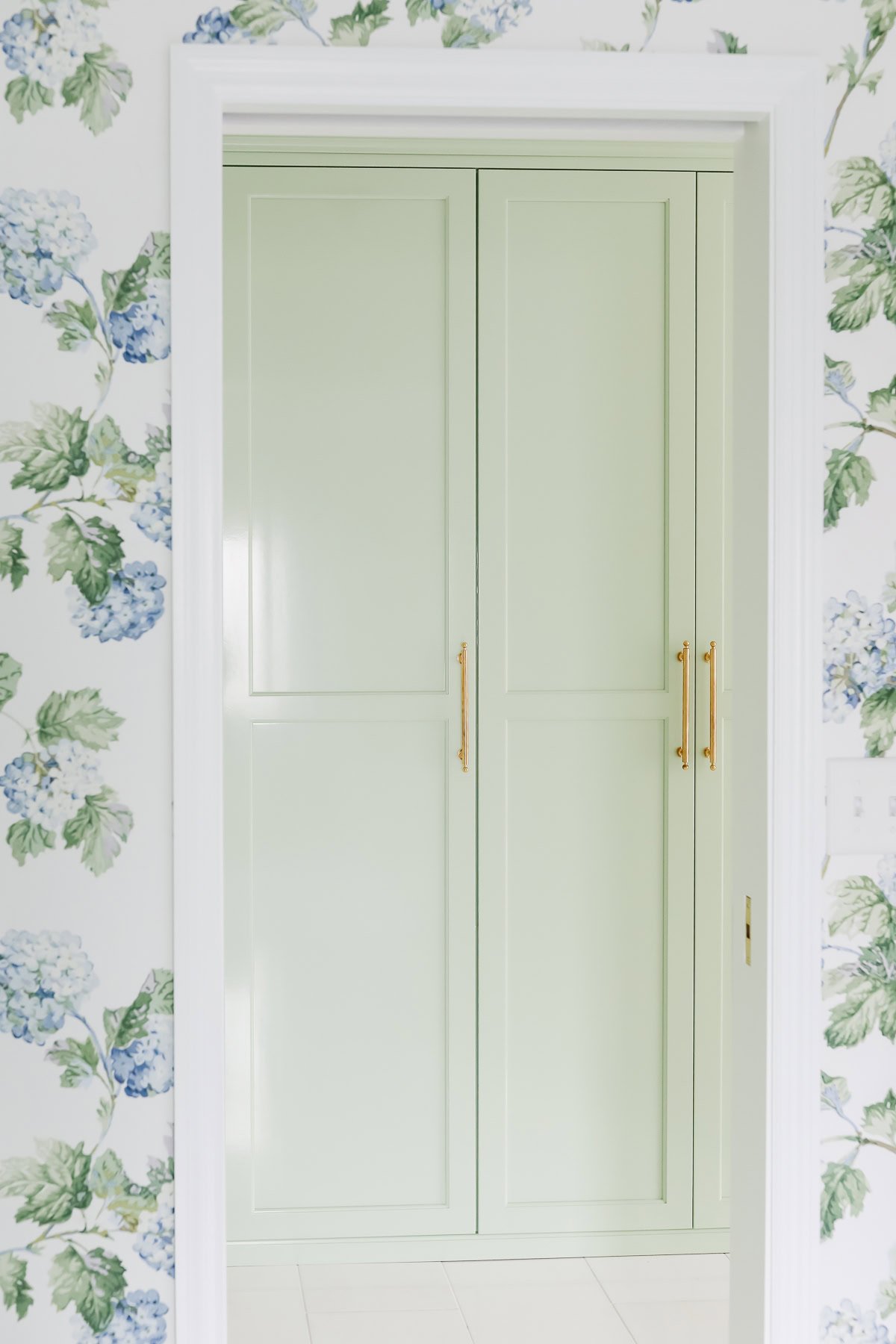 Light green double doors with gold handles, framed by white trim, set against a floral wallpaper with blue and green hydrangea patterns.