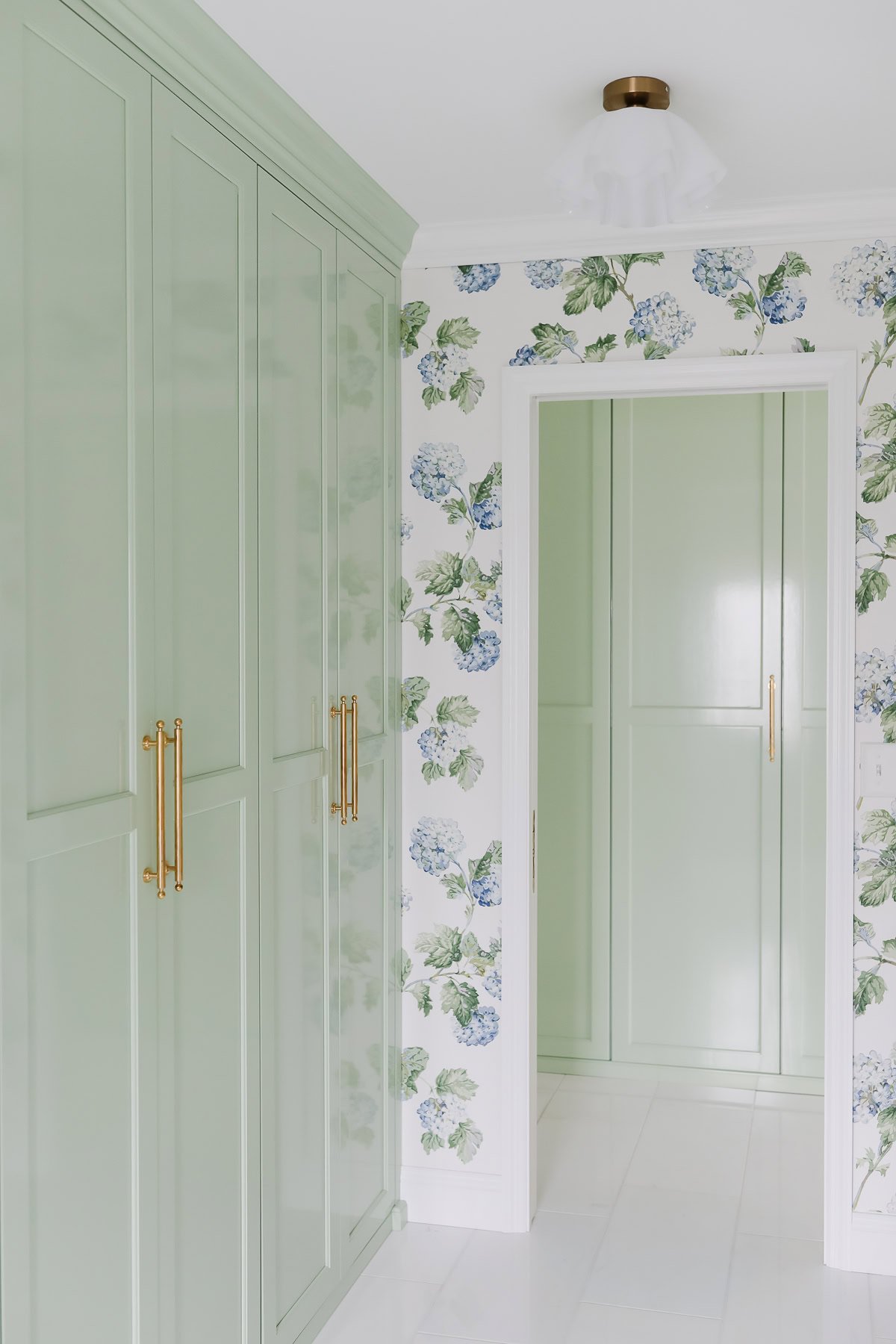 A hallway with light green cabinets, gold handles, white flooring, and floral wallpaper featuring blue and green hydrangeas.