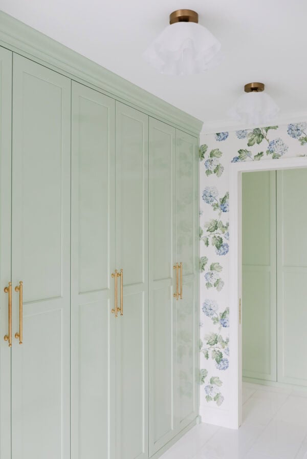 A hallway with pale green built-in cabinets featuring gold handles, white tile flooring, floral wallpaper, and two white ceiling lights with brass bases.