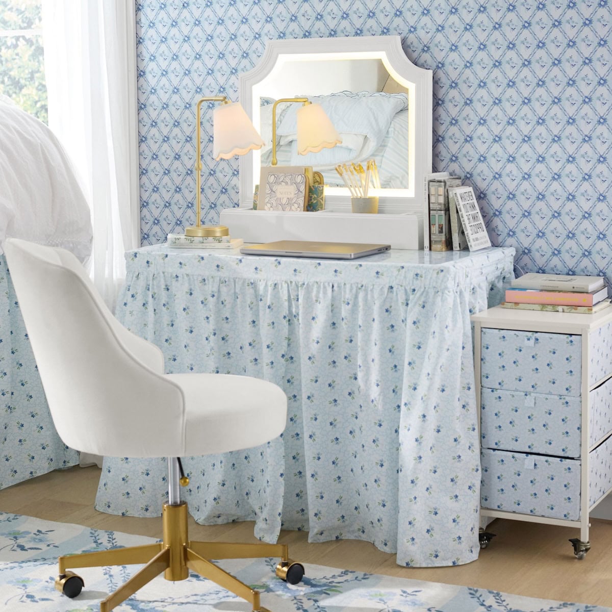 A vanity desk with a mirror, gold desk lamps, and stationary items sits against a blue floral wallpapered wall, paired with a white swivel chair and matching drawer unit.