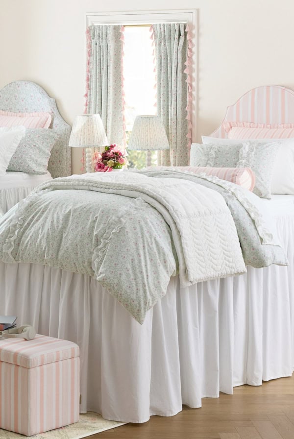 A neatly made bed with floral bedding and white ruffled bed skirt stands between two bedside tables, each with a lamp. Matching curtains cover a window behind the bed.