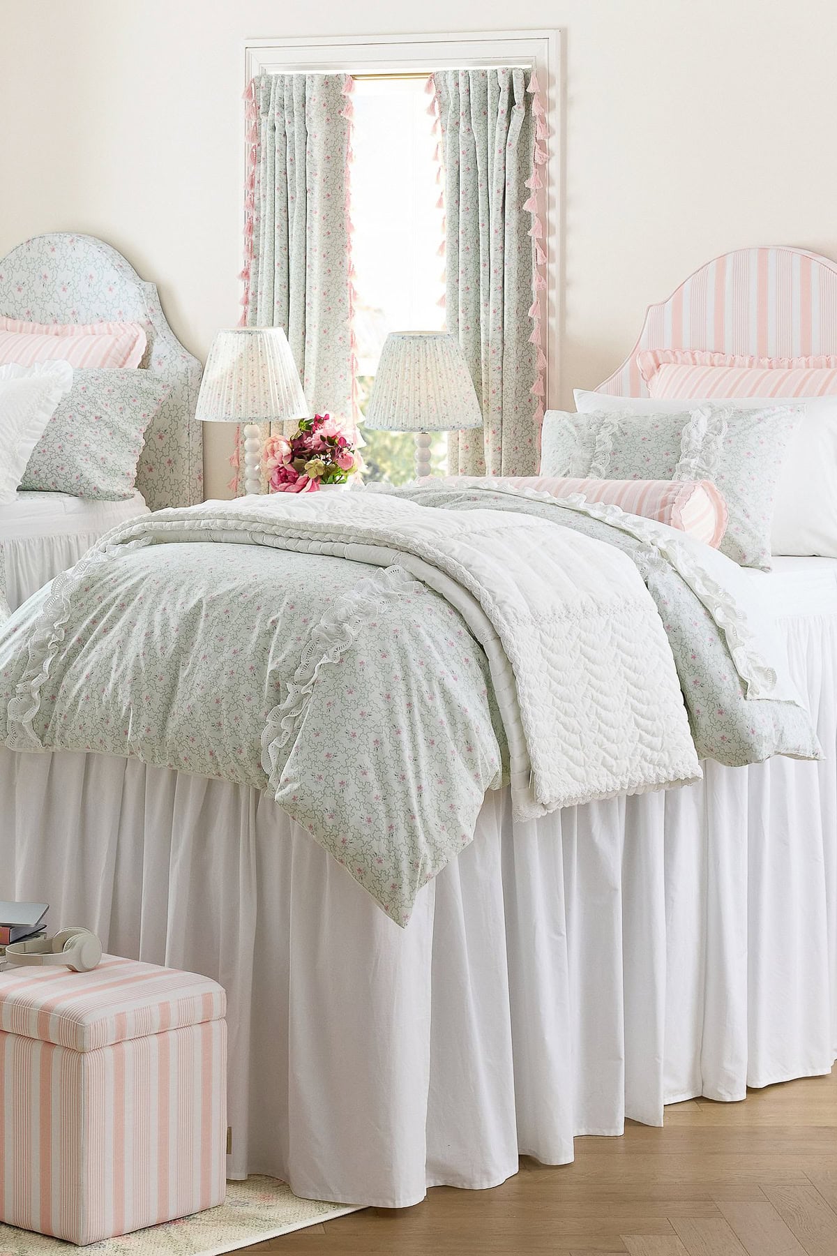 A neatly made bed with floral bedding and white ruffled bed skirt stands between two bedside tables, each with a lamp. Matching curtains cover a window behind the bed.